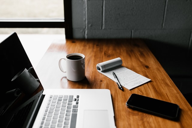 An office desktop next to a window. Atop the desk is a MacBook, a mug of black coffee, a mobile phone, and a notepad and pen.